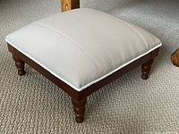 Full view of square beige upholstered footstool on carpet