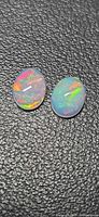 Both opal cabochons side by side on black textured surface showing play-of-color