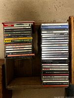 Two stacks of approximately 45 music CDs in wooden crate