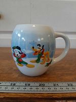Front view showing Mickey and Pluto graphics on curved mug with ruler for scale