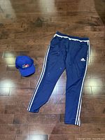 Full view of navy Adidas track pants with white stripes and blue baseball cap