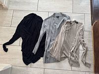 Three sweaters laid flat: black draped, grey open-front, beige button-front