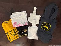 All six tops laid out on floor: yellow St. Clair College T-shirt, white Orlando T-shirt, black diamond graphic T-shirt, two white ribbed tank tops, grey John Deere hoodie