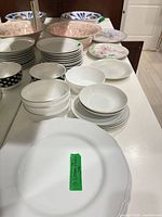 Assorted stacks of plates and bowls including scalloped white dinner plate, salad plates, polka-dot bowls, pink speckled serving bowl, blue/green patterned serving plates, floral plate.
