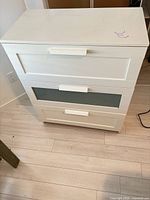Front view of white 3-drawer chest, showing paneled fronts and frosted glass insert