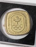 Front of square medallion showing maple leaf, Olympic rings, bilingual inscription, laurel wreath border