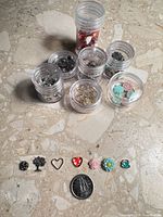 Group of seven clear plastic jars filled with assorted miniature charms, several sample charms lined in front next to Canadian dime for scale