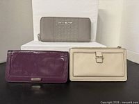Three wallets front view: grey Michael Kors zip-around studded wallet, purple Cole Haan wallet, beige Kate Spade wristlet wallet