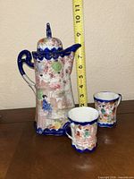 Front view of pot with lid and two cups beside measuring tape indicating height