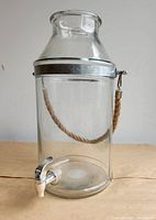 Front view of glass beverage dispenser showing body, neck, metal collar, rope handle and spigot