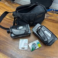 Lot overview showing two Sony camcorders, Sony carrying case, sealed Fujifilm Hi8 tape, sealed Canon memory card