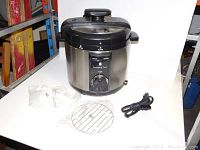 Full front view of Wolfgang Puck electric pressure cooker with power cord, metal rack, and plastic accessory on white surface.