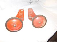Pair of vintage training wheels with orange rims and attached mounting brackets, showing wear and use.
