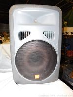 Front view of large JBL speaker cabinet showing grey plastic housing, vent openings and metal grille over the woofer cone.
