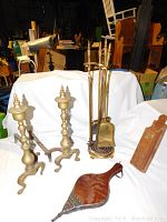 Two photos showing a pair of brass andirons, a brass fireplace toolset with stand, and two wooden fireplace accessories (bellows and wooden paddle) arranged on a white backdrop.