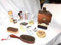 Photo showing all shoe shine kit items arranged around wooden shoe shine box with brass latch including brushes, tins, and bottles of polishes and conditioners