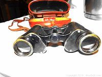 Photo of the vintage black binoculars with brass accents in front of the open brown leather carrying case on a white surface.