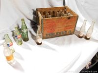 Wooden crate with multiple vintage soda bottles around it on white cloth background.
