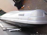 Side view of silver Thule car top storage unit with black base lying on ground.