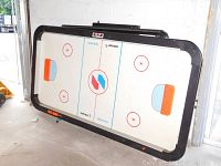 Full view of the air hockey table placed upright showing the playing surface, goals, and black frame with electronic scoring unit.