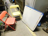 Set of two children's folding chairs, one yellow and one red, shown folded and standing next to the whiteboard table.