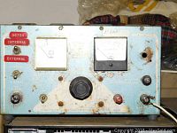 Front view of the vintage AMP machine showing two analog meters, control knob, toggle switches, and connectors. Blue metal casing with rust spots.