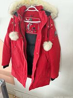 Full view of red parka on hanger showing hood fur trim, pompoms, front closure, overall style