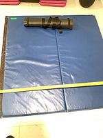 Blue crash mat unfolded with measuring tape showing size; black rolled exercise mat resting on top
