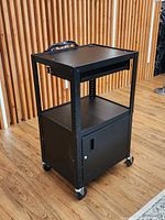 Three-quarter view of black steel AV cart showing handle, shelves, cabinet, casters