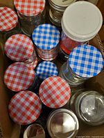 Cardboard box containing multiple clear glass jam jars with gingham lids in red, blue and white patterns plus a few silver lids