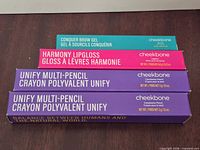 Stacked unopened boxes: Conquer brow gel, Harmony lip gloss, two Unify multi-pencils