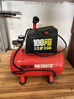 Side view of red Central Pneumatic compressor showing 100 PSI label, tank, handle and hose