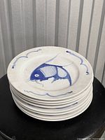 Stack of eight white ceramic plates showing blue koi fish design and wave border.