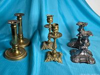 All six candle holders grouped: brass column pair, ornate brass square-base pair, silverplate ornate base pair