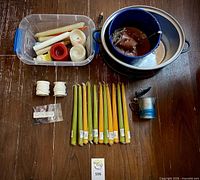 All candle making components laid out including candles, dye chips, wicks, cooker, pots, ladle, plastic bin