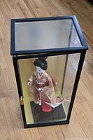 Front view of black-framed glass case containing geisha doll