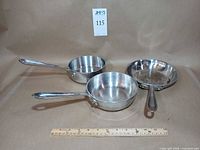 Three stainless steel All-Clad pans with ruler for scale