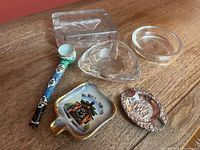 Group shot of all six smoking accessories arranged on tabletop: three glass ashtrays (rectangular, triangular, round), ceramic cuckoo clock ashtray, silver-plate figural ashtray, painted pipe