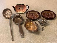 Group shot showing ladle, tea strainer spoon, pink tea bag holder, two brown pottery dishes, Birks dish and teapot-shaped strainer