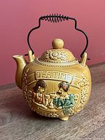Front view showing embossed couple, TEA TIME banner, floral motifs, handle, lid and feet