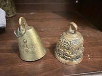 Two brass bells side by side: grape-embossed cowbell and ornate Christian bell