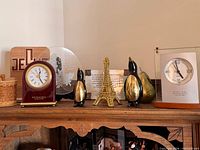 Top shelf showing two desk clocks, Eiffel Tower, penguin pair, pear, woven jar, wood Jeep plaque and round Japanese glass plaque