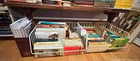 Two white wire baskets filled with assorted paperback and hardcover books on floor under shelf