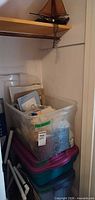 Closet view showing clear tote filled with yarn, cross-stitch supplies, embroidery frames, additional tote beneath, and sailboat on shelf