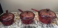 Three Visions cranberry glass saucepans/casseroles with lids displayed on countertop