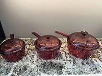 Three cranberry glass saucepans with lids displayed side by side
