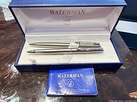 Open presentation case displaying two Waterman silver ballpoint pens with booklet