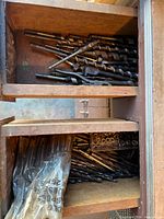 Open wooden cabinet shelves containing auger bits on top, twist drill bits in bag and loose on lower shelf, small metal stops visible