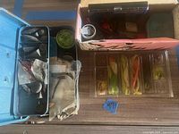 Overview showing blister pack of soft lures, tackle box tray with hooks, sinkers, spool of line, and cardboard box containing mixed lures and bobbers
