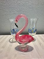 Pink glass flamingo figurine displayed in front of two hurricane glasses